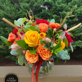 Round glass vase of yellow, orange, and red roses with small yellow mums and berries, tied with an orange ribbon