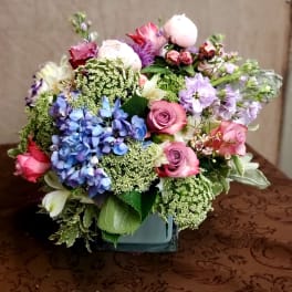 Mixed bouquet of pink, blue, and lavender flowers in a square vase