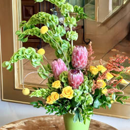 Tall floral arrangement with pink protea, yellow roses, and green blooms in a vase