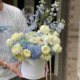 White hatbox arrangement with cream roses, blue hydrangeas, and pale blue delphinium