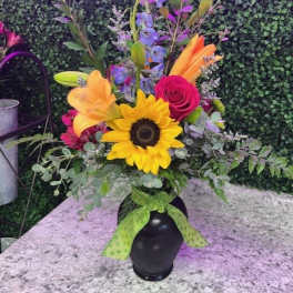 Tall mixed bouquet with sunflower, lilies, rose, and blue delphinium in a black vase with green bow.