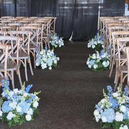 Low blue and white flower arrangements line a wedding aisle between wooden chairs and a black curtain backdrop.