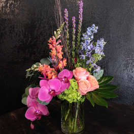 Tall modern arrangement with pink orchids, peach roses, blue delphinium and green hydrangea in a glass vase