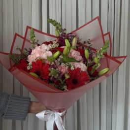Handheld bouquet of red and pink flowers wrapped in pink paper with a white ribbon
