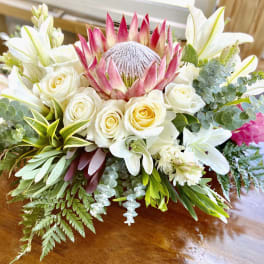 Low arrangement of white roses, lilies, and a pink protea with lush foliage on a wooden table