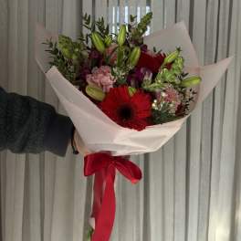 Handheld bouquet of red gerbera daisies, pink carnations, and lily buds wrapped in pale pink paper