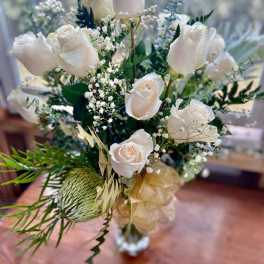 Tall arrangement of white roses with baby's breath, a green protea, and gold ribbon in a clear vase.