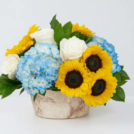 Blue hydrangeas, white roses, and yellow sunflowers in a rustic container