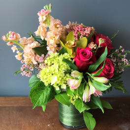 Mixed bouquet of red roses, green hydrangea, orchids, and peach blooms in a green-wrapped glass vase