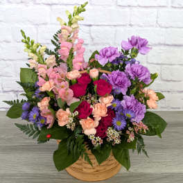 Basket arrangement of pink snapdragons, purple carnations, peach spray carnations, and red roses.
