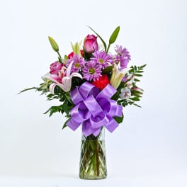 Bouquet of pink lilies, roses, and purple daisies in a glass vase with a purple bow
