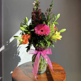 Tall mixed bouquet with pink gerbera daisy, orange roses, lilies and snapdragons in a clear vase with pink bow