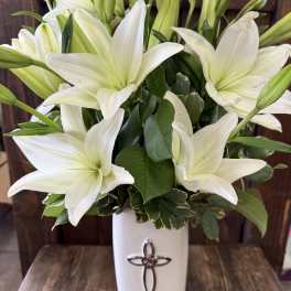 White lilies arranged in a tall white vase with a cross emblem