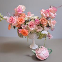 Pink and peach rose arrangement in a white pedestal vase with a rose-shaped teapot