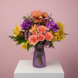 Peach, yellow, and purple mixed flower arrangement in a purple vase on a white pedestal