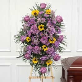 Tall standing spray of lavender roses and yellow daisies on an easel beside a wooden casket.