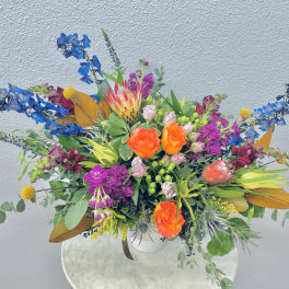 Bright mixed bouquet with orange roses, blue flowers, and pink blooms in a white vase