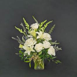 White roses, lilies, and snapdragons arranged in a clear glass vase against a dark background