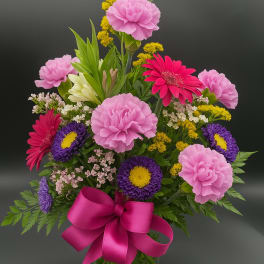 Mixed bouquet of pink carnations, gerbera daisies, and purple asters in a glass vase with a pink bow