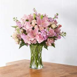 Pink lilies, roses, and mixed flowers arranged in a clear glass vase on a wooden table