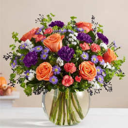 Mixed bouquet of orange roses, purple carnations, and small daisies in a round clear glass vase