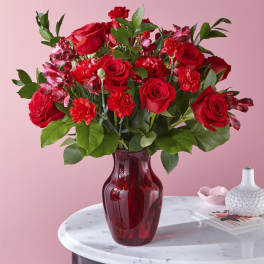 Arrangement of red roses and carnations in a red glass vase on a marble table.