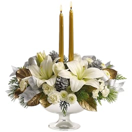 White lilies and roses arranged in a glass compote with two gold taper candles