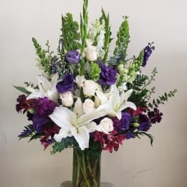 Tall floral arrangement with white lilies, roses, and purple blooms in a glass vase