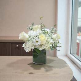 Low arrangement of white and cream roses, hydrangeas, and daisies in a clear glass cylinder vase