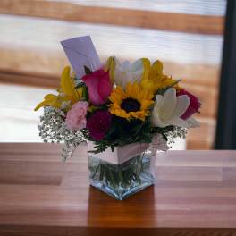 Mixed bouquet in a clear square vase with sunflowers, lilies, and roses
