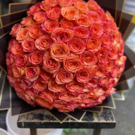 Large spherical bouquet of orange and pink roses wrapped in dark fan-folded paper.