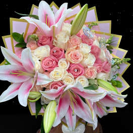 Pink lilies and cream and pink roses arranged in a white vase with decorative wrap and butterfly accents