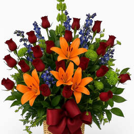 Basket arrangement of orange lilies, red roses, blue delphinium and green blooms with a red bow