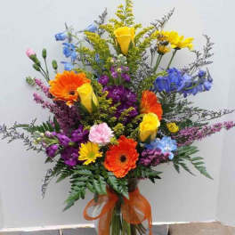 Tall mixed arrangement of bright yellow roses, orange gerberas, and purple and blue flowers in a glass vase