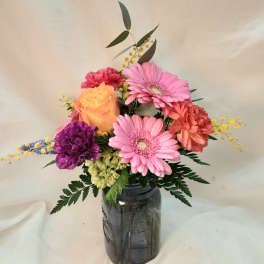 Colorful mixed arrangement of pink gerbera daisies, orange carnations, and yellow rose in a tinted glass jar