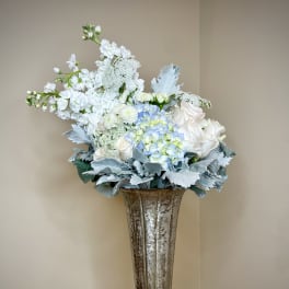 Tall white and soft blue flower arrangement in a metallic trumpet vase on a round draped table.