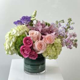 Short arrangement of pink roses, orchids, and green hydrangeas in a dark green glass cylinder vase