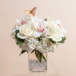 White floral arrangement in a clear square vase with a butterfly topper
