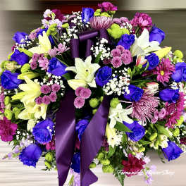 Large oval arrangement of purple, blue, white, and pink flowers with a purple ribbon bow