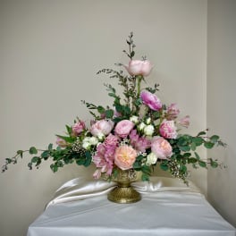 Asymmetrical arrangement of pink and peach roses and ranunculus in a gold pedestal vase with trailing greenery.