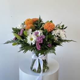 Mixed bouquet of orange, white, and purple flowers in a glass vase with a white ribbon