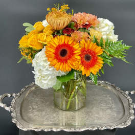 Bright mixed bouquet in a glass vase with orange gerberas, yellow roses, and white hydrangeas.