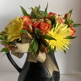 Orange roses and yellow chrysanthemums in a black pitcher vase with a gold ribbon