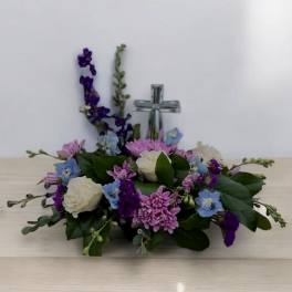 Low arrangement of white roses, lavender mums, and blue blooms with a glass cross in the center
