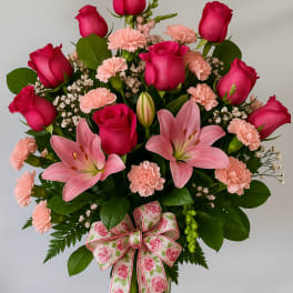 Tall arrangement of hot pink roses, pink lilies, and light pink carnations in a clear vase with a floral bow.