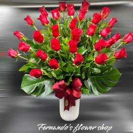Tall arrangement of red roses in a white vase with large glossy leaves and a red ribbon bow