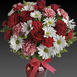 Red roses, white daisies, and pink carnations in a red vase with a polka-dot ribbon