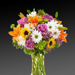 Mixed bouquet of roses, lilies, daisies, and sunflowers in a clear glass vase