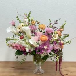 Asymmetrical pastel arrangement with calla lilies, ranunculus, orchids, and mixed blooms in a clear glass vase
