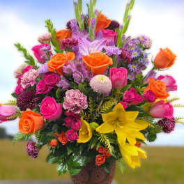 Tall mixed arrangement of pink and orange roses, yellow lilies, and purple accents in a brown pot